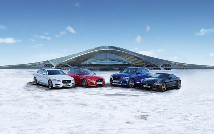 Four Jaguar cars parked on a snowy surface in front of a modern architectural building under a clear blue sky. The cars from left to right include a white Jaguar XE, a red Jaguar XF with license plate S1 JAG, a blue Jaguar F-PACE with license plate S1 JAG, and a black Jaguar F-TYPE with license plate S1 JAG. The image showcases new cars, highlighting sleek design and luxury, suitable for advertising used cars and new cars.