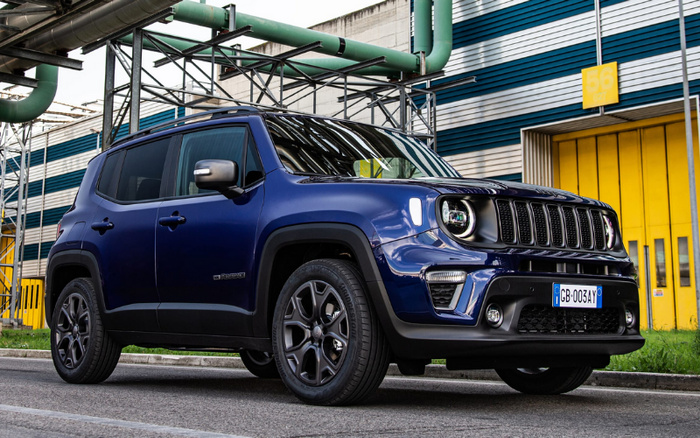 Blue Jeep Renegade SUV parked on an urban street in front of a building with yellow doors and blue striped walls, featuring green industrial pipes overhead. The vehicle has black alloy wheels and a European license plate reading CB-003AY. The image showcases a new car model, suitable for promotions involving used cars and new cars.
