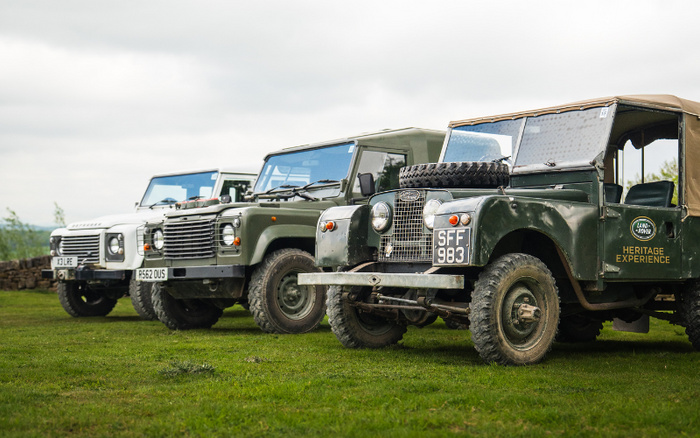 Line of vintage Land Rover vehicles parked on grass, including a green Land Rover with a spare tyre on the bonnet and license plate SFF 983 labelled 'Heritage Experience', followed by two other Land Rovers with license plates P562 OUS and XJ LFE, showcasing classic used cars and new cars in a rural outdoor setting under an overcast sky.