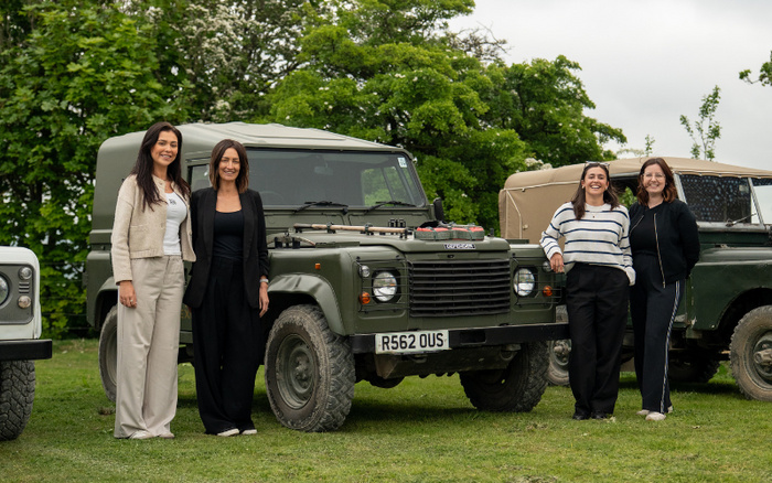 Four women standing beside classic green Land Rover vehicles on a grassy area with trees in the background, showcasing used cars. The vehicles have licence plates including R562 OUS, indicating these are used cars. The setting suggests a display or gathering emphasising the appeal of both used cars and potentially new cars in the off-road or classic vehicle category.