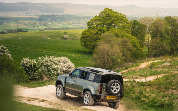 Land Rover Defender 110 with UK registration plate KM74 EYP driving on a dirt road in a green countryside landscape with rolling hills and trees in the background, illustrating rugged terrain suitable for both used cars and new cars.