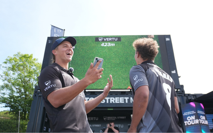 Two young men wearing black and grey Vertu branded sports shirts stand outdoors in front of a large digital screen displaying 'VERTU 423m'. One man is holding a smartphone and appears to be excited or celebrating. The backdrop includes signage with the text 'BRISTOL STREET MOTORS' and 'ON TOUR', indicating a promotional event likely related to used cars and new cars.
