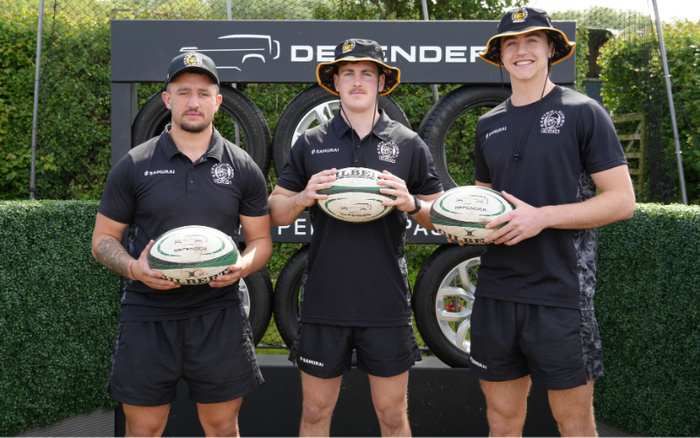Three men wearing black Samurai sportswear and hats holding rugby balls, standing in front of a black Defender tyre and parts display with visible text 'DEFENDER'. The image promotes used cars and new cars through the backdrop featuring car tyres and the Defender brand.