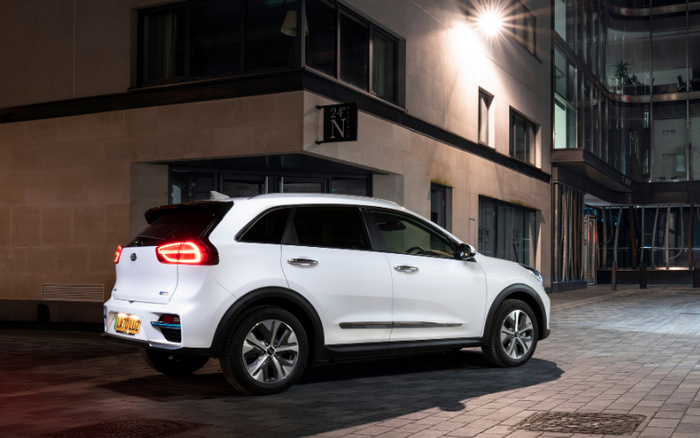 White Kia electric SUV parked on a city street at night in front of modern buildings, with illuminated rear lights and a visible yellow UK license plate, symbolising both used cars and new cars available in urban environments.
