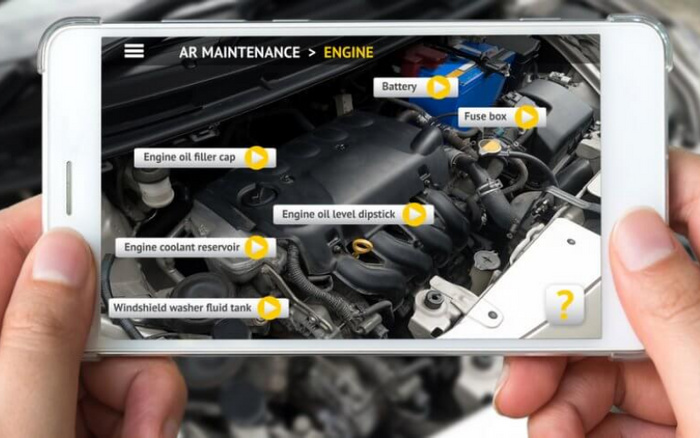 Smartphone displaying augmented reality (AR) maintenance interface focused on a car engine, with labelled components including battery, fuse box, engine oil filler cap, engine oil level dipstick, engine coolant reservoir, and windshield washer fluid tank. The screen shows navigation path at top reading 'AR MAINTENANCE > ENGINE'. Hands holding the phone capture the engine compartment of a used or new car.