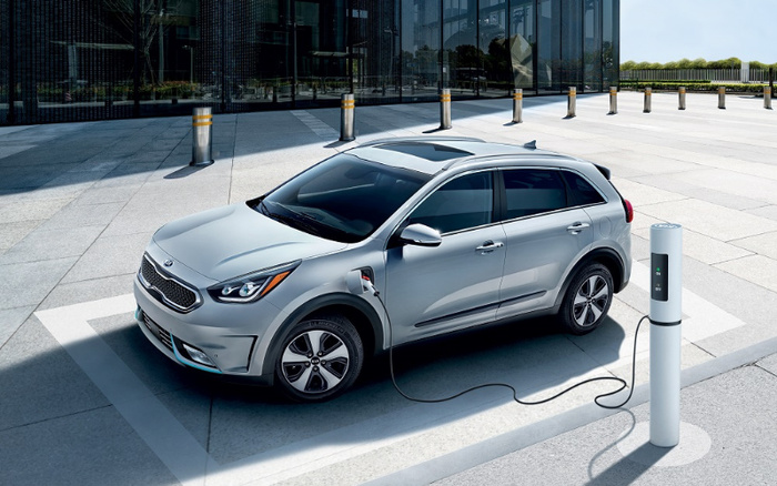 Silver electric Kia Niro plugged into a white charging station in a modern urban setting with reflective glass building walls in the background. The vehicle is parked on a paved area with clear sunlight, showcasing a sleek design and tinted windows. Keywords: used cars, new cars.
