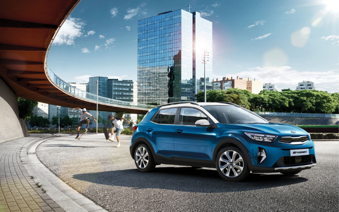 Blue Kia Stonic parked on the side of a city street under a curved bridge with modern glass buildings and greenery in the background, with two people skateboarding nearby. The image highlights a new car model, suitable for showcasing both new cars and used cars in an urban environment.