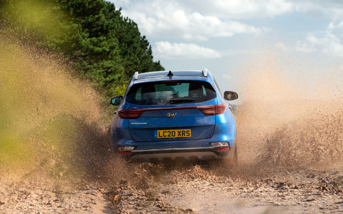Rear view of a blue Kia Sportage with UK registration plate LC20 XRS driving at speed on a muddy dirt road, splashing mud on both sides, under a partly cloudy sky with green trees in the background, illustrating a rugged, off-road driving experience with used cars and new cars.