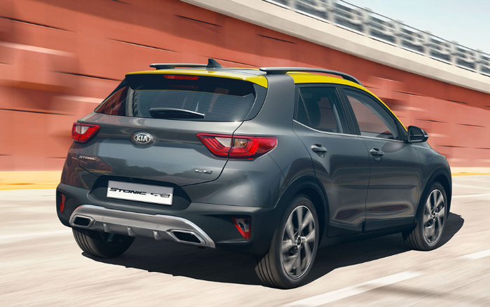 Rear view of a grey Kia Stonic hybrid car with yellow roof accents driving on a road next to a red wall, highlighting a modern new car model suitable for buyers interested in used cars and new cars.