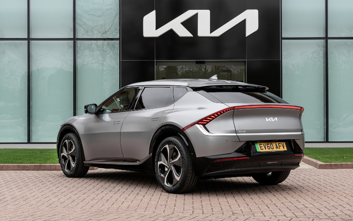 Rear side view of a silver Kia EV6 electric car parked outside a Kia dealership with large black and glass panel exterior. The car features distinctive rear light design and UK license plate EV60 AFV. The setting highlights the availability of used cars and new cars at the dealership.