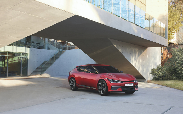 Red Kia EV6 GT-Line electric car parked outside a modern building with large concrete structures and glass panels. The car is positioned on a smooth concrete surface near stairs and greenery. Optimised for used cars and new cars.
