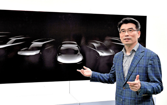 Man in a blue checked suit and glasses standing next to a large image of conceptual car silhouettes on a black background, possibly presenting or explaining new car designs. The image hints at innovation and features sleek outlines of multiple vehicles, relevant for used cars and new cars markets.