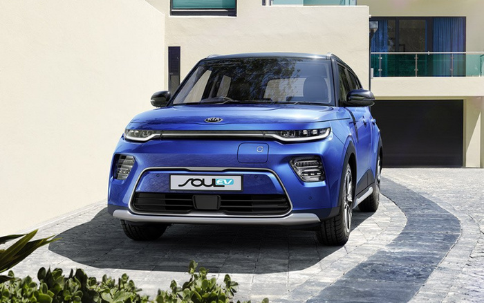 Front view of a blue Kia Soul EV electric vehicle parked on a paved driveway in front of a modern house with cream walls and large windows, showcasing the car's sleek design, LED headlights, and distinctive grille, ideal for buyers interested in used cars and new cars.