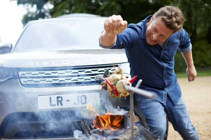 Man seasoning a whole chicken roasting on a spit over an open fire, with a grey Land Rover Discovery parked in the background, displaying the front grille, 'DISCOVERY' badge, and a license plate reading 'LR - JQ', representing used cars and new cars.