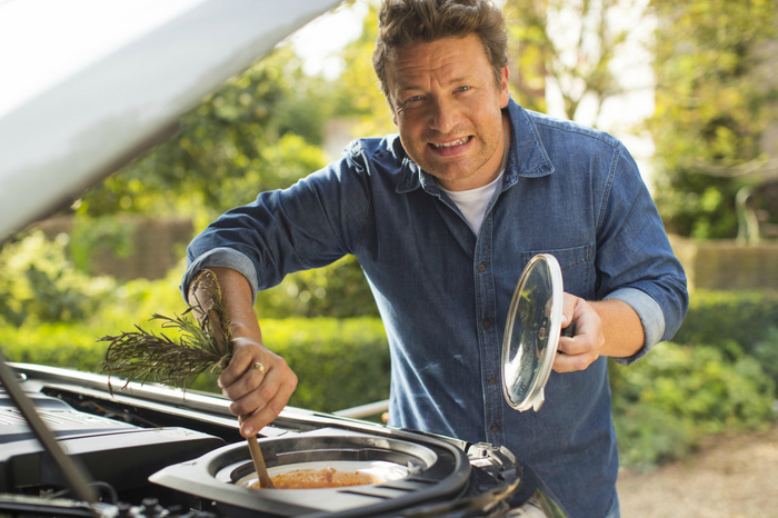 Man in a denim shirt stirring a pot with herbs under the open bonnet of a car, outdoors in a garden setting, humourously using the car engine bay as a slow cooker. Conceptual image combining cooking and car themes, relevant for used cars and new cars marketing with a creative twist.