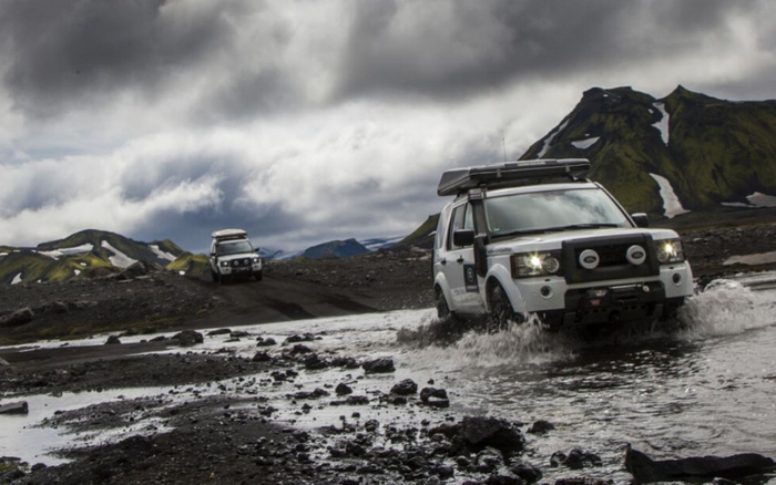 Two white off-road vehicles driving through a rugged, rocky landscape with shallow water crossing, under a dramatic cloudy sky with mountainous terrain in the background. The vehicles appear capable and sturdy, suitable for adventurous travel, representing robust used cars and new cars designed for challenging environments.