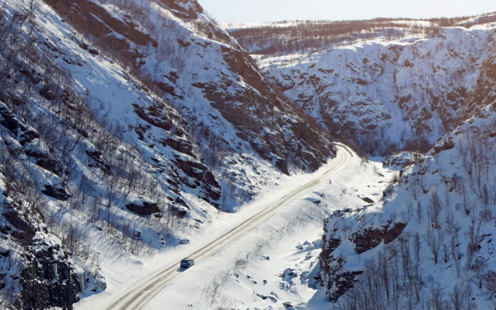 Snow-covered mountain road winding through a rocky, mountainous landscape with sparse trees and bushes, featuring a single blue car driving on the road in winter conditions. The scene highlights the rugged terrain and the challenges of driving in snowy weather, relevant to used cars and new cars in harsh environments.
