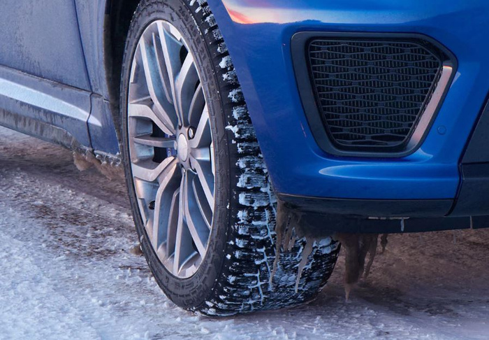 Close-up of a blue car's front wheel and bumper on a snowy surface. The tyre tread is visible with traces of snow and ice, highlighting winter driving conditions. The car features a modern alloy wheel design. The image relates to used cars and new cars in winter conditions.