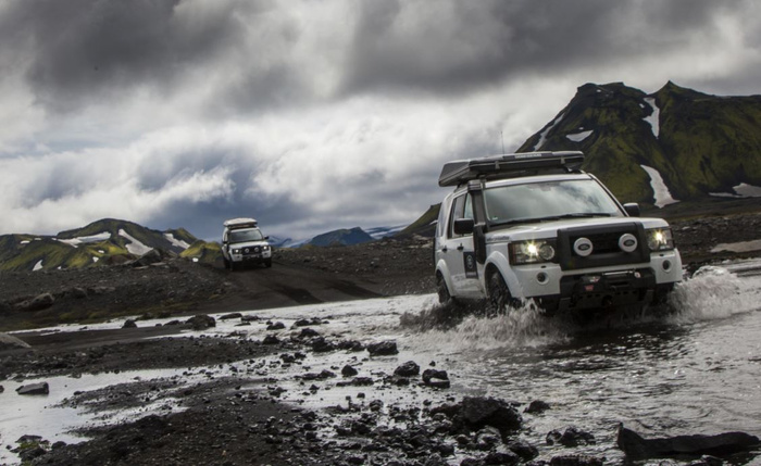 Two white off-road vehicles driving through rocky terrain and shallow water under a cloudy sky with mountainous landscape in background, showcasing rugged used cars and new cars suitable for adventurous off-road driving.
