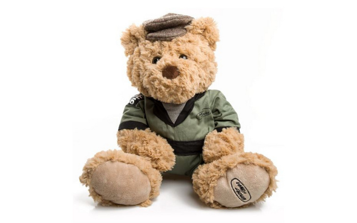 A plush teddy bear wearing a green outfit and a brown flat cap sits against a white background. The bear's outfit includes a small embroidered logo on the left arm and chest reading 'Arnold Clark.' The sole of the bear's left foot is embroidered with the text 'Arnold Clark.' The image is focused on the teddy bear and contains no references to used cars or new cars.