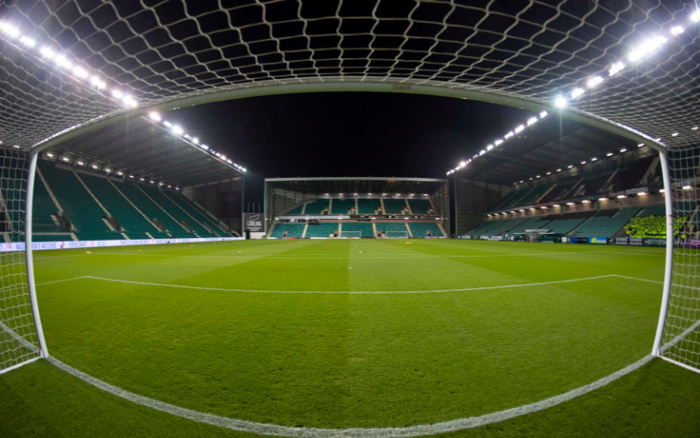View from inside a football goal looking out onto a well-lit, empty stadium pitch at night with green seats in the stands and a digital advertising board displaying text about used cars and new cars.
