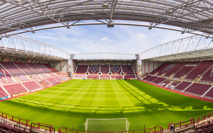 Panoramic view of an empty football stadium with well-maintained green pitch and maroon seating stands under a steel roof structure, no text related to used cars or new cars visible in the image