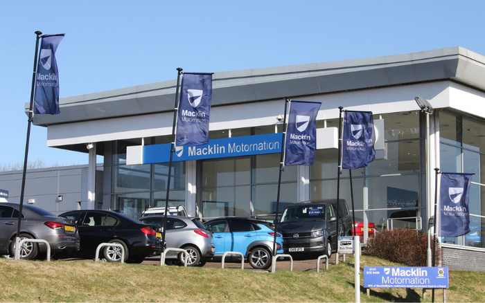 Exterior view of Macklin Motornation car dealership showing a modern glass-fronted building with five Macklin Motornation flags. Several cars, including black, silver, blue, and grey vehicles, are parked outside on a grassy frontage. A blue sign in the foreground displays contact details for Macklin Motornation, noting '9141 879 9499' and a statement that '98% of Macklin Motornation customers would recommend them to a friend'. The dealership offers both used cars and new cars for sale.