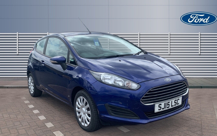 Blue Ford Fiesta hatchback car parked in front of a modern building with horizontal metal panels. The car has a UK registration plate reading SJ15 LSC and features a prominent front grille with the Ford logo above it. The image includes the Ford logo in the top right corner. This car is suitable for both used cars and new cars searches.
