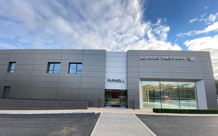 Exterior view of the Farnell Land Rover dealership building under a partly cloudy sky, featuring a modern grey façade with large glass windows and entrance doors. The building displays the Land Rover logo and name prominently, representing a location for buying new cars and used cars.