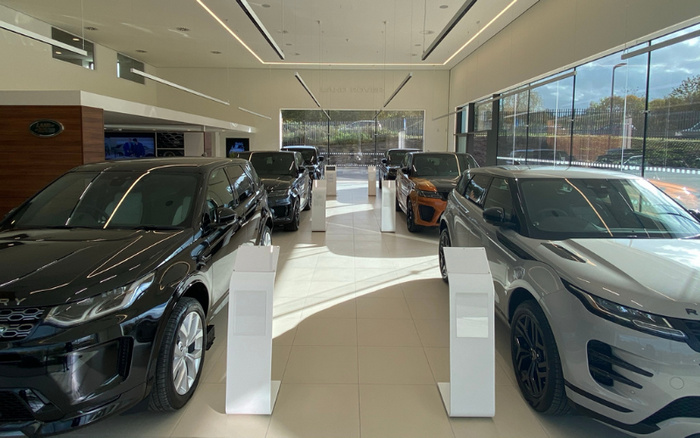 Bright, modern Land Rover showroom displaying an array of new cars including black, grey, and orange models on shiny beige tile flooring, with large windows allowing natural light to fill the space, and white information stands placed beside each vehicle showcasing details about the used cars and new cars available for sale.