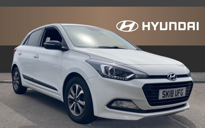 White Hyundai hatchback car with registration plate SK18 UFG parked on a concrete surface in front of a brown and black wall featuring the Hyundai logo and the word 'HYUNDAI' in white letters, showcasing a used car in a dealership setting for used cars and new cars.