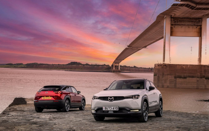 Two Mazda cars parked on a cobblestone surface near a river under a large bridge at sunset, featuring a white Mazda with license plate BU73 AYD facing forward and a red Mazda with license plate BU73 AXT facing away. The scene highlights both used cars and new cars with a scenic background of a colourful sky and distant hills.