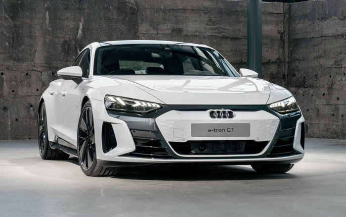 White Audi e-tron GT electric sports car parked indoors against a rough concrete wall, showcasing sleek headlights, distinctive grille with Audi rings, and aerodynamic design, ideal model for new cars and used cars listings.