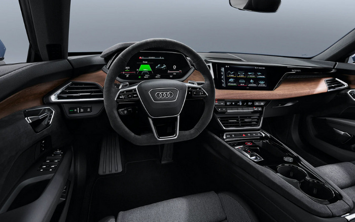 Interior view of a modern Audi car featuring a digital dashboard behind a multi-function steering wheel with the Audi logo at the centre, a sleek infotainment screen integrated into a wooden dashboard panel, climate control buttons below the screen, and black fabric seats, highlighting premium design and technology suitable for both new cars and used cars buyers.