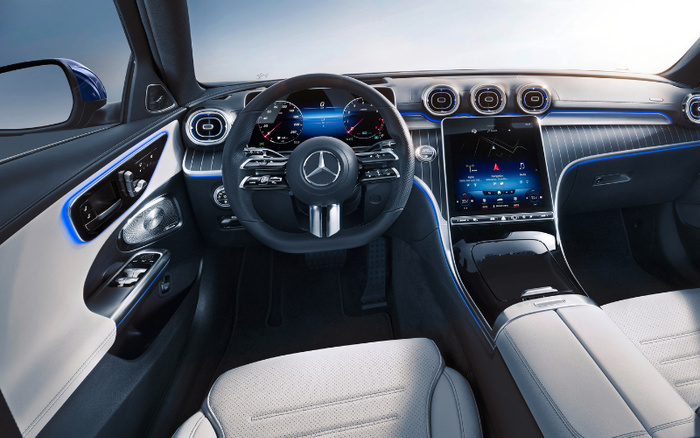 Interior of a luxury Mercedes car showing the steering wheel with Mercedes logo, digital instrument cluster displaying speedometer and tachometer, and central touchscreen with navigation map. The dashboard and door panels feature ambient blue lighting and sophisticated design with leather and wood trim. The image highlights the modern, high-tech cockpit environment typical of new cars, suitable for marketing used cars or new cars in an upscale segment.