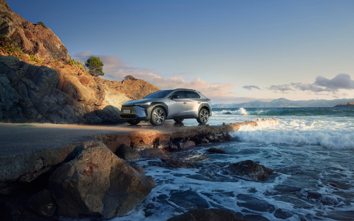 Silver SUV parked on a narrow concrete pier by the rocky seaside with waves crashing below, under a clear blue sky with distant mountains and clouds, showcasing a new car model ideal for both used cars and new cars buyers.