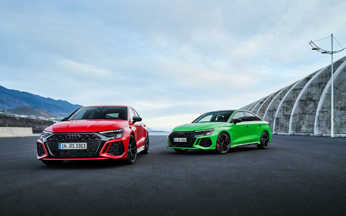Two high-performance Audi cars parked on a wide asphalt surface near a large modern curved concrete structure with mountainous landscape in the background. The car on the left is a red Audi RS 3301 with sporty styling, and the car on the right is a green Audi RS 3302. Both cars feature sleek designs with black alloy wheels and aggressive front grilles. The scene highlights premium, sporty vehicles suitable for those interested in used cars or new cars.