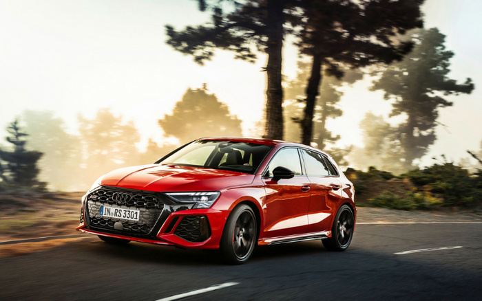 A red Audi RS3 hatchback driving on a road surrounded by trees in soft sunlight. The car features sporty black alloy wheels, a honeycomb front grille, and LED headlights. The front license plate reads 'IN RS 3301'. This image represents new cars available in the market and is suitable for illustrating used cars and new cars options.