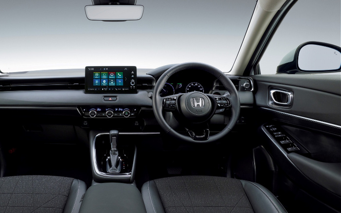 Interior view of a modern Honda car showing the dashboard, steering wheel with Honda logo, central touchscreen display with menu icons, gear shift, air conditioning controls, and black fabric seats. The image highlights the sleek design of a new car, suitable for used cars or new cars listings.