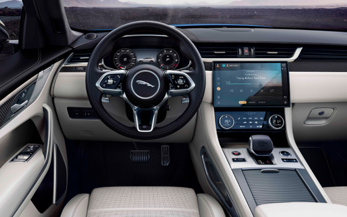 Interior view of a luxury Jaguar car showcasing the steering wheel with the Jaguar logo, digital instrument cluster displaying driving information, centre console featuring a large touchscreen infotainment system playing the song 'Young Before Your Time' by Echosmith, dual climate control settings, modern gear selector, and beige leather seats, ideal for those interested in used cars and new cars.