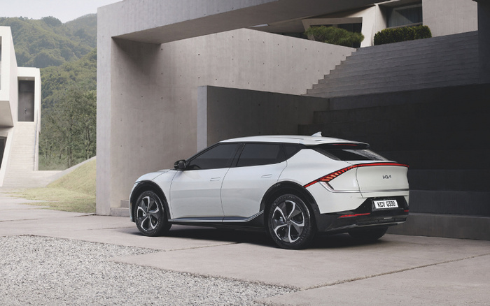 White Kia electric vehicle parked outside a modern concrete house with minimalist architectural design and outdoor steps; car features sleek aerodynamic body, tinted windows, and distinctive rear light design, with license plate reading KCV 0309; background shows green hills and gravel driveway, representing new cars and used cars in a stylish setting.