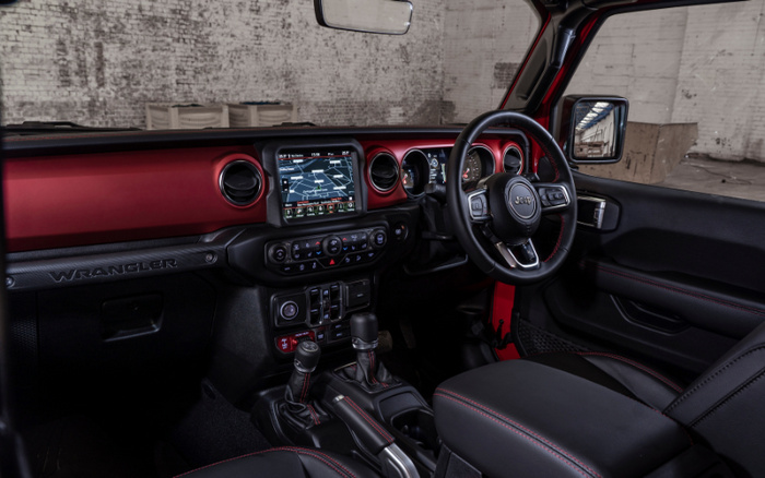 Interior view of a Jeep Wrangler showcasing the dashboard, steering wheel with Jeep logo, touchscreen navigation system, gear shifters, and black leather seats with red stitching, highlighting the modern features of new cars and appealing design for buyers interested in used cars or new cars.