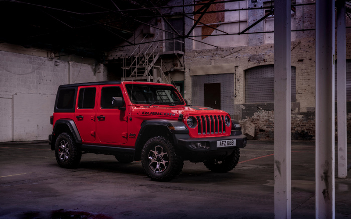 Red Jeep Rubicon parked inside a dimly lit industrial warehouse with concrete floors and tall metal pillars. The vehicle has a black roof, rugged off-road tires, and a visible front license plate reading AFZ 668. The interior of the warehouse features exposed brick walls, a staircase, and large windows with some natural light entering. This image is relevant for used cars and new cars in an industrial setting.