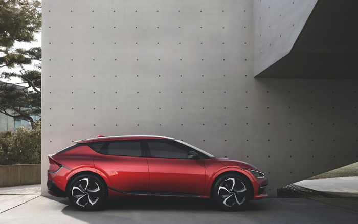 Sleek red modern hatchback parked beside a minimalist concrete wall with small regularly spaced circular indentations. The car features aerodynamic design with black window trims and stylish alloy wheels. Surrounded by greenery on one side and open space on the other, suitable for advertising used cars or new cars.