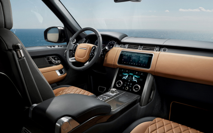 Interior of a luxury car showing the driver's seat, steering wheel, and dashboard with advanced digital displays, surrounded by ocean view through the windows. The car features quilted tan leather seats, black and tan dashboard with carbon fibre centre console, and dual touchscreen controls displaying climate and media information. Ideal for buyers searching for used cars or new cars with premium interior features.