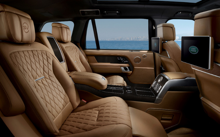 Luxury car interior with brown quilted leather seats featuring embossed 'SV' logos on the headrests, spacious rear seating area with centre console, wood and carbon fibre trim, panoramic sunroof, and dual rear entertainment screens displaying the Land Rover logo, set against a backdrop of a sea view through the windows, showcasing the premium design of used cars and new cars.