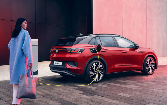 Red Volkswagen ID.4 GTX electric car being charged at a modern charging station next to a building with black and pink walls. A woman in a blue coat carrying a grey tote bag walks towards the car. The car's license plate reads 'ID.4 GTX'. The scene combines elements of new cars and electric vehicle technology, suitable for advertising used cars and new cars.