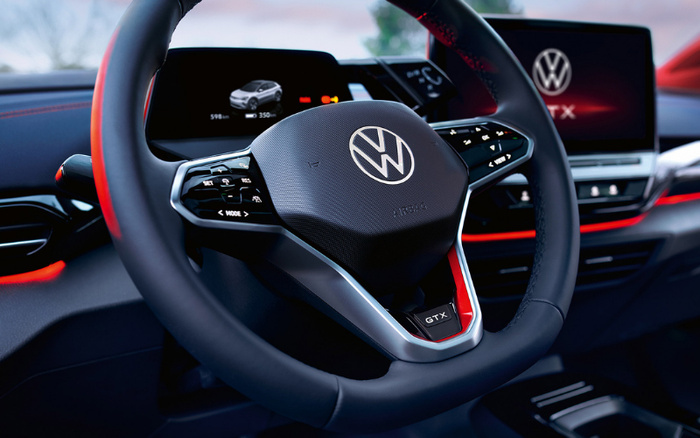 Close-up of a Volkswagen GTX car interior showing the steering wheel with the VW logo and GTX badge. The dashboard displays a digital instrument cluster with car information such as range and mileage, alongside a central infotainment screen featuring the Volkswagen logo and GTX branding. Interior accents include red stitching and ambient lighting.