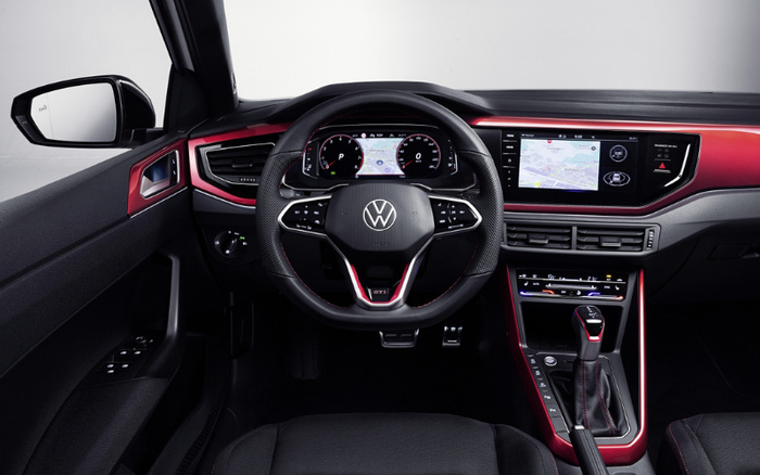 Interior view of a Volkswagen car dashboard and steering wheel featuring a digital instrument cluster displaying navigation maps, speedometer, and gear indicator. The steering wheel has the VW logo with GTI branding. The dashboard includes a touchscreen infotainment system also showing navigation, air vents, climate control panel, and gear shift lever with red accents. Side mirror and part of the driver’s seat and door with control buttons are visible. Suitable for used cars and new cars sales or reviews.