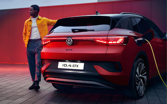 Rear view of a red Volkswagen ID.4 GTX electric car charging at a station, with a man in a yellow jacket and grey trousers standing beside it against a red wall background. The car features distinctive LED tail lights and the model badge 'GTX' below the VW logo and license plate reading 'ID.4 GTX'. This image represents new cars and is relevant for showcasing both used cars and new cars in automotive contexts.